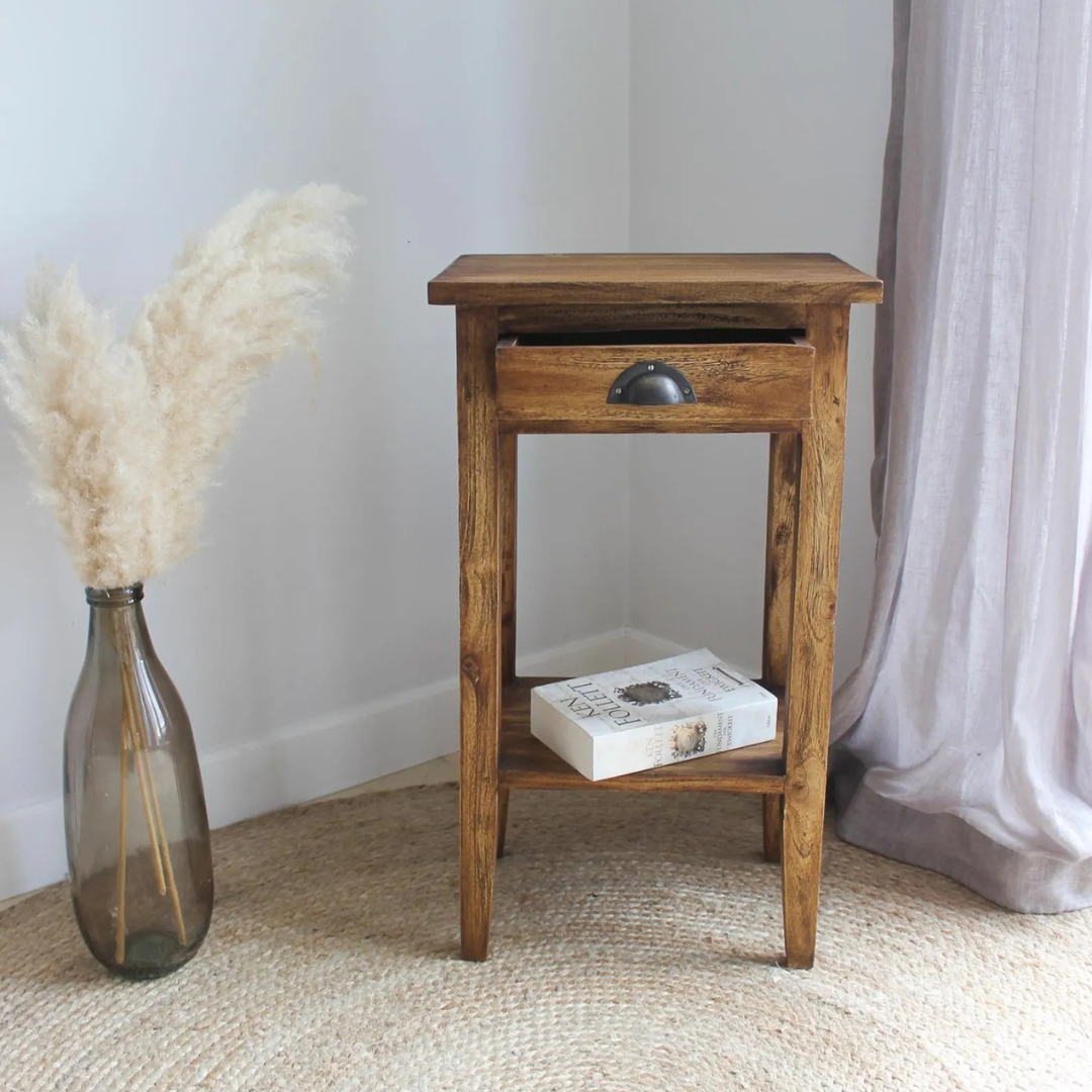 A rustic wooden nightstand with a single drawer and open lower shelf, styled in a room setting next to a glass vase with pampas grass and a book on a woven rug.