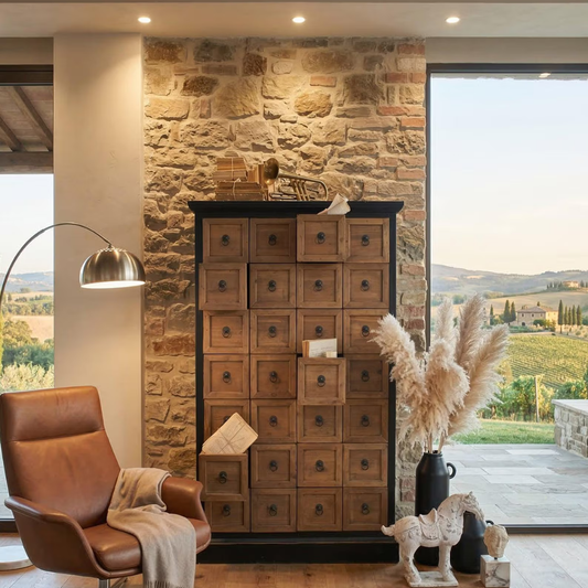 A tall wooden cabinet with multiple small drawer fronts and round metal knobs, placed in a rustic interior with a stone wall, large windows, a brown leather chair, and decorative pampas grass.