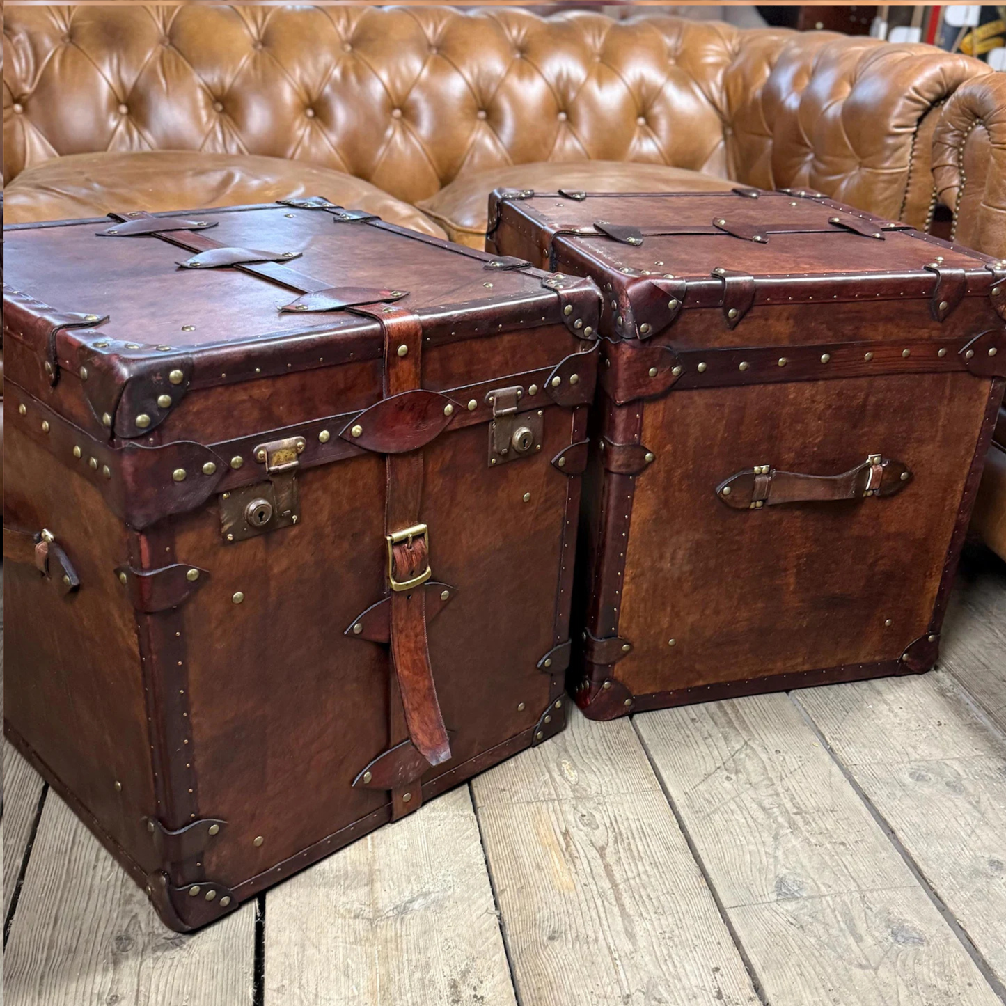 Two vintage-style brown leather trunks on a wooden floor with a brown leather sofa in the background.