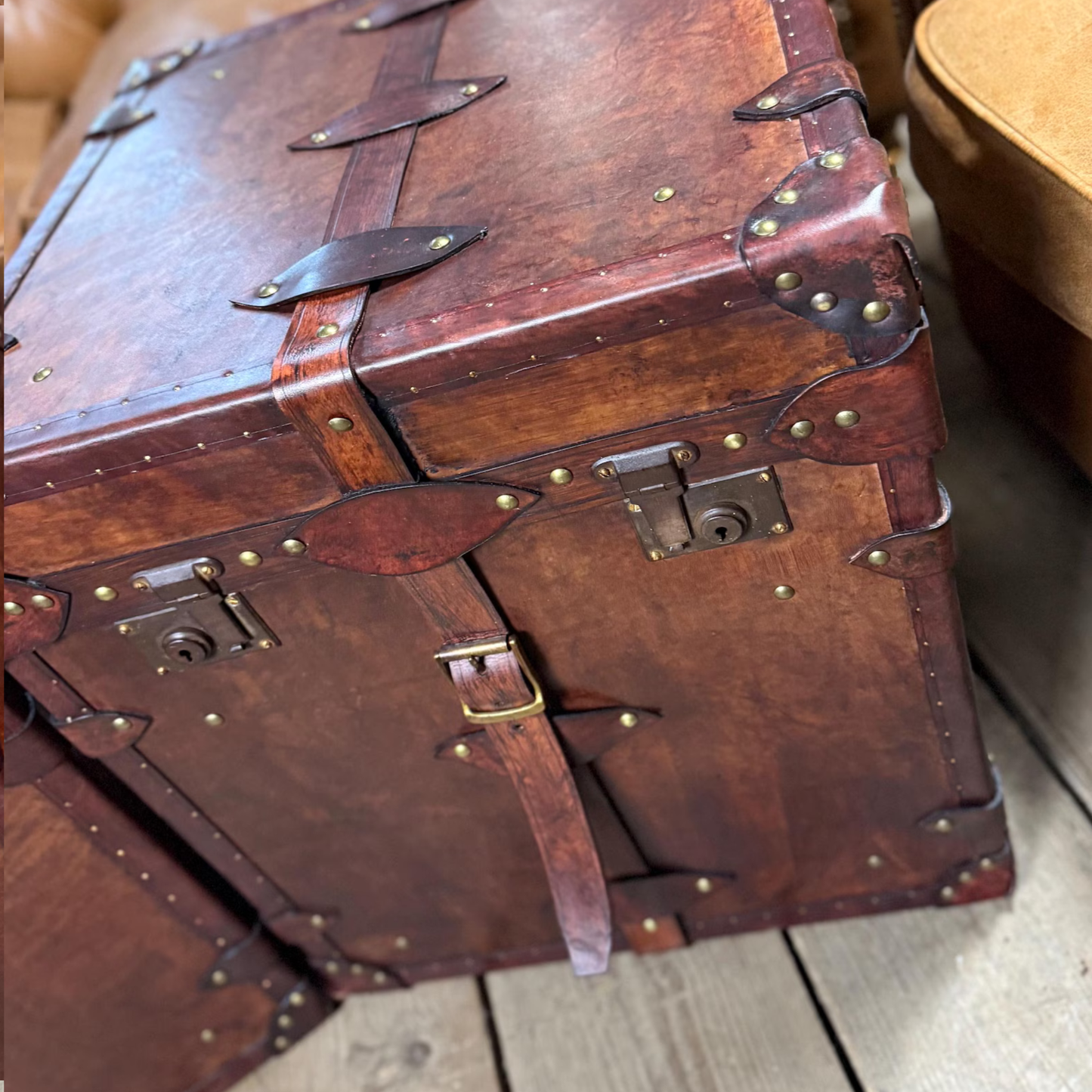 Vintage brown leather trunk with brass studs and buckles on a wooden floor.