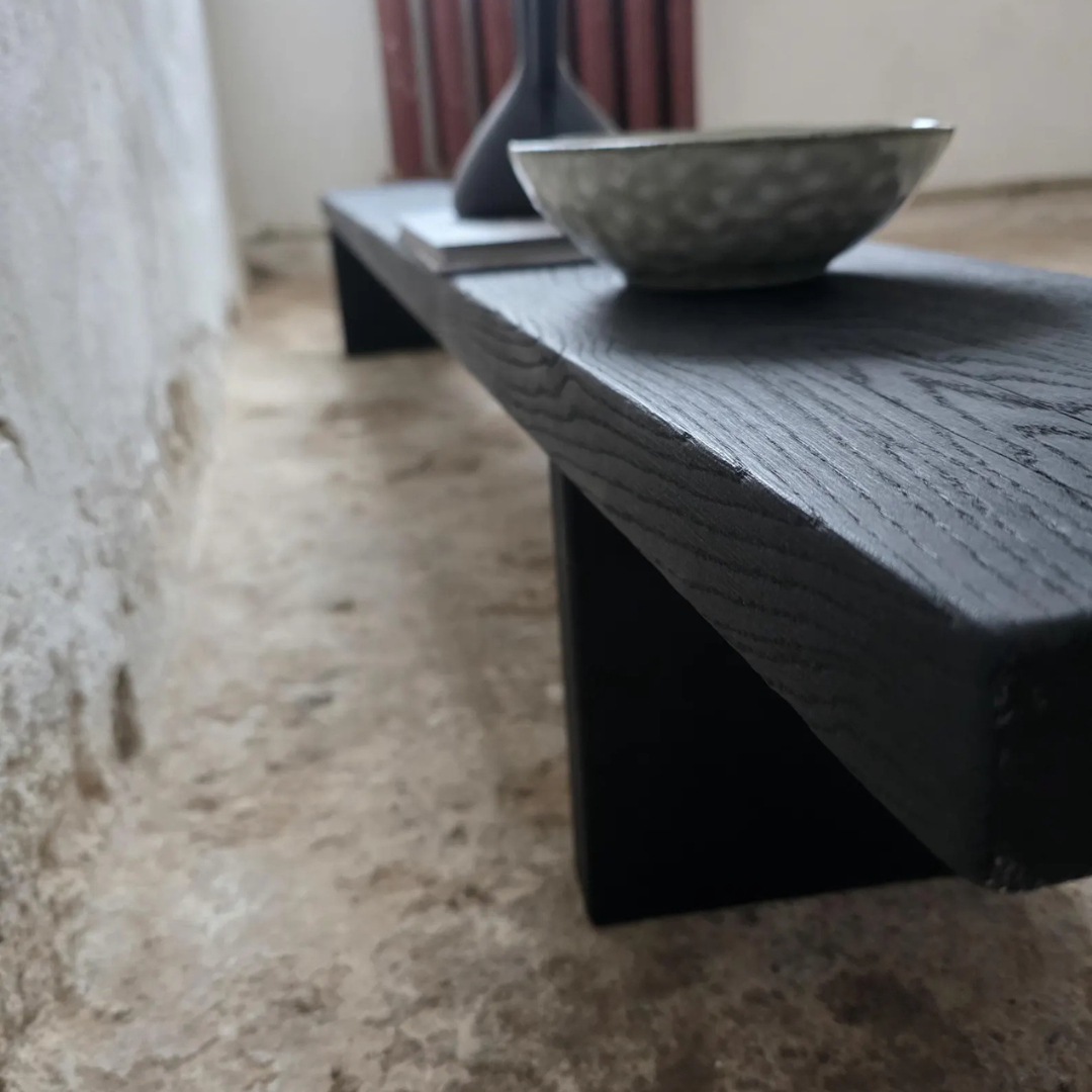 A low black wooden bench with a thick plank top and block-style legs, shown in a rustic room setting with a textured concrete floor and a gray bowl on top.