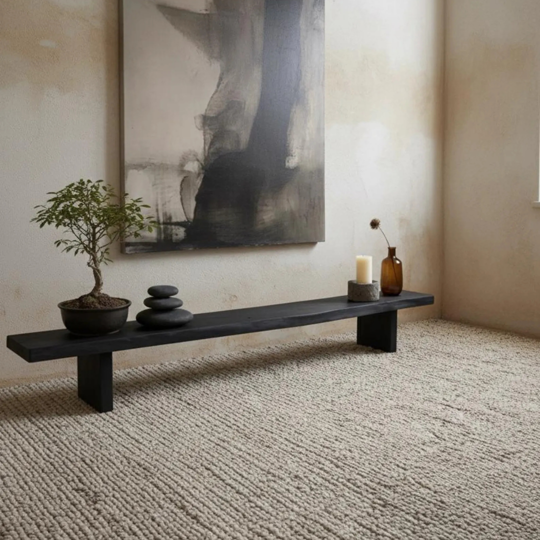 A long black wooden bench with block legs sits on a textured beige rug in a minimalist room. Decorative items on the bench include a potted bonsai tree, stacked black stones, a white candle, and an amber glass bottle.
