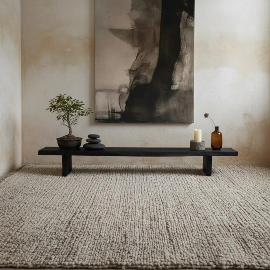 A long black wooden bench styled in a minimalist room with a textured beige rug, a potted bonsai tree, stacked stones, a candle, and a small amber glass vase, set against a warm plaster wall with a large abstract black and gray painting.
