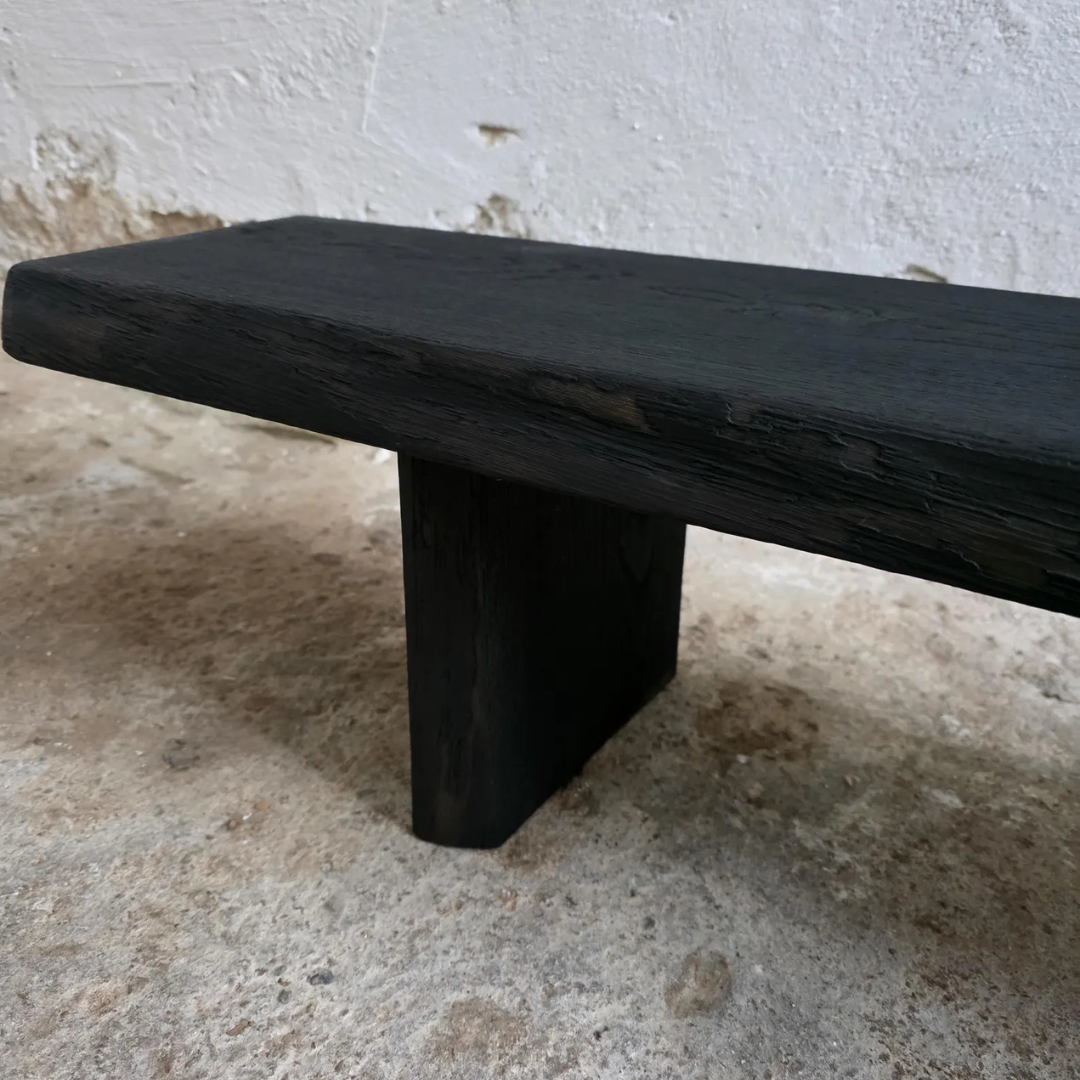 A rustic black wooden bench with a thick rectangular seat and a single block-style leg, photographed against a textured off-white wall and concrete floor.