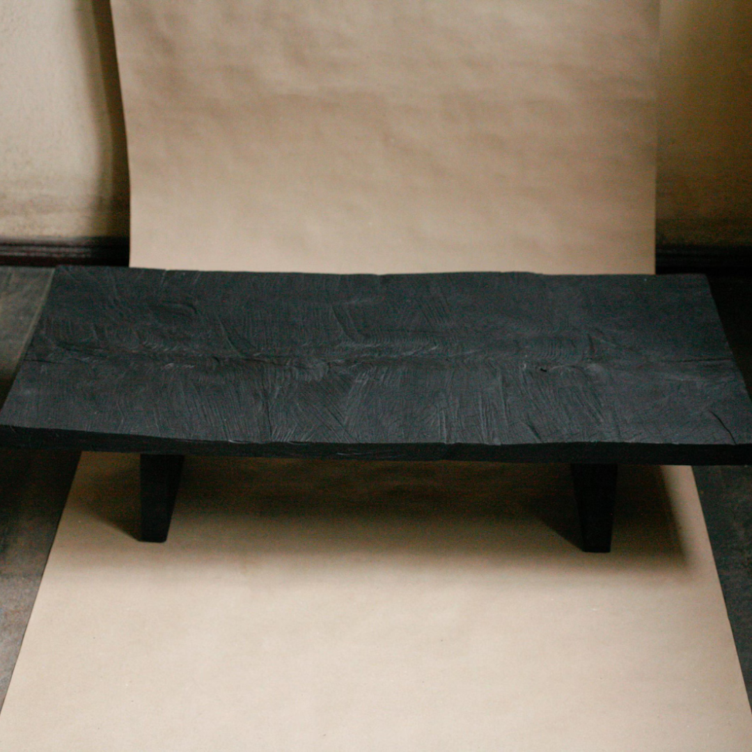 A low, rectangular black bench or stool with a heavily textured, cracked surface and short legs, photographed against a backdrop of beige paper sheets.