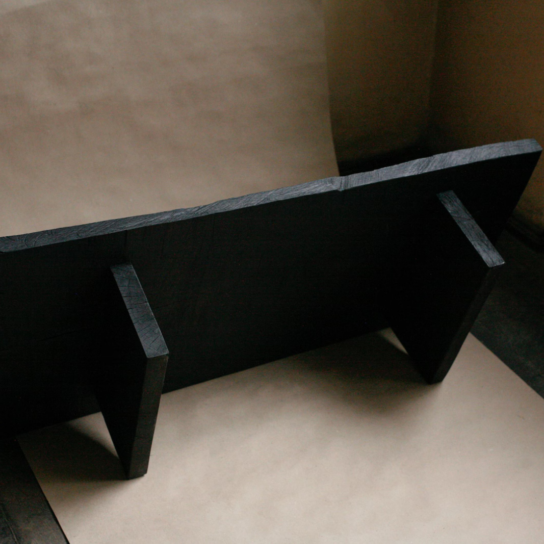 A dark charcoal gray stone bench with a thick, rustic flat top and two short legs, photographed in a minimalist studio setting with beige paper backgrounds.