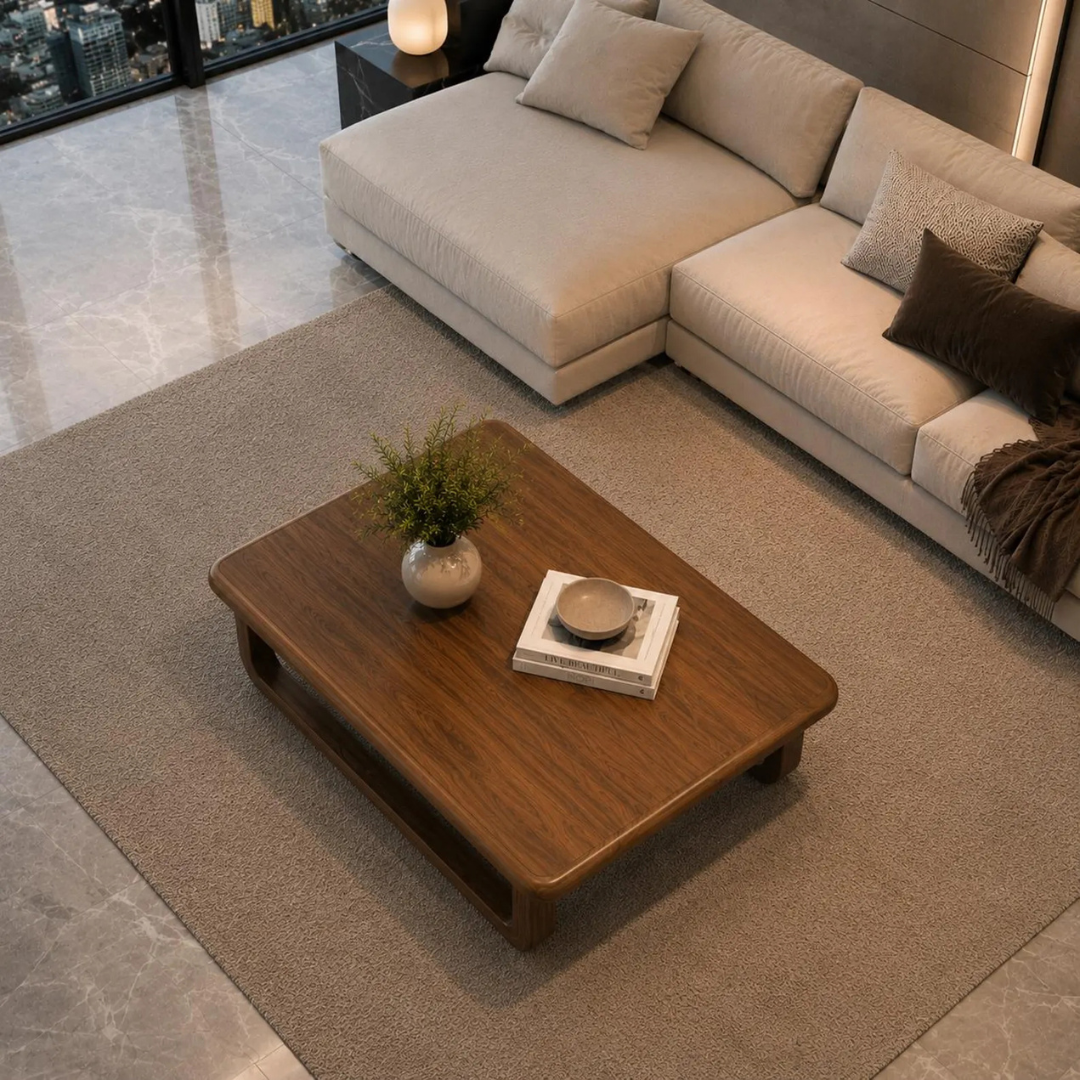 A modern living room scene featuring a low, rectangular wooden coffee table with a warm medium-brown finish, styled with a small potted plant and books. The table is placed on a large beige area rug, with a light beige sectional sofa and dark brown accent pillows in the background.