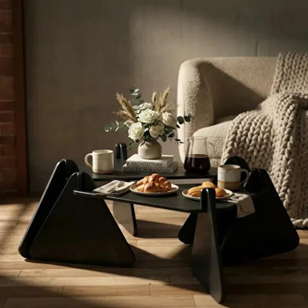 A modern black coffee table with sculptural angular legs in a warm, sunlit living room. The table is styled with a vase of white flowers, books, mugs, and plates of pastries, positioned in front of a cream-colored armchair with a chunky knit throw blanket.