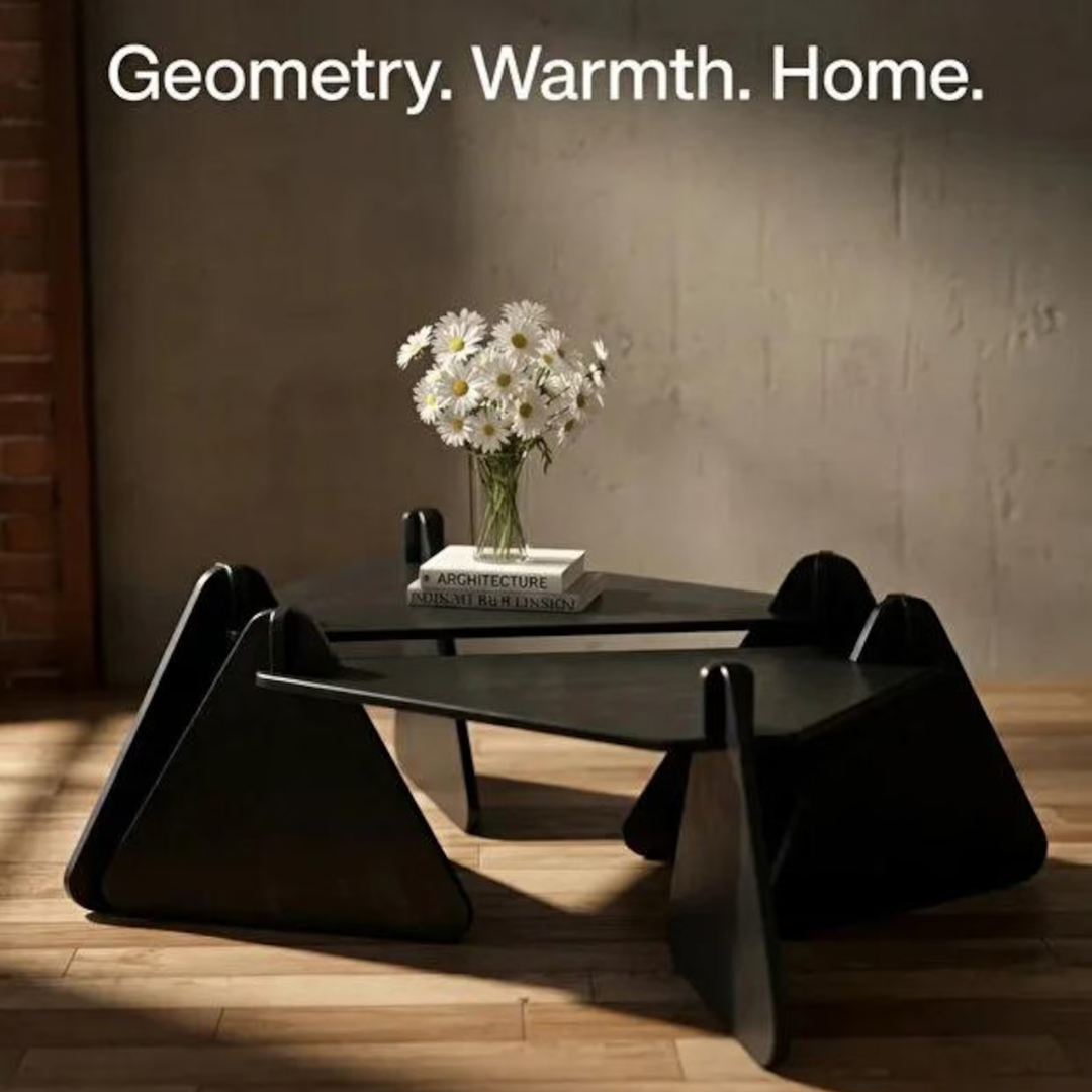 A modern black geometric coffee table with angular triangular legs, styled with a vase of white daisies and a book on a dark tabletop, set in a warmly lit room with wood flooring and a textured wall.