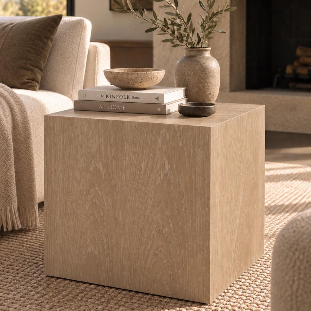 A square light oak wood end table styled in a cozy living room setting, holding a stack of books, a ceramic bowl, and a vase with olive branches.