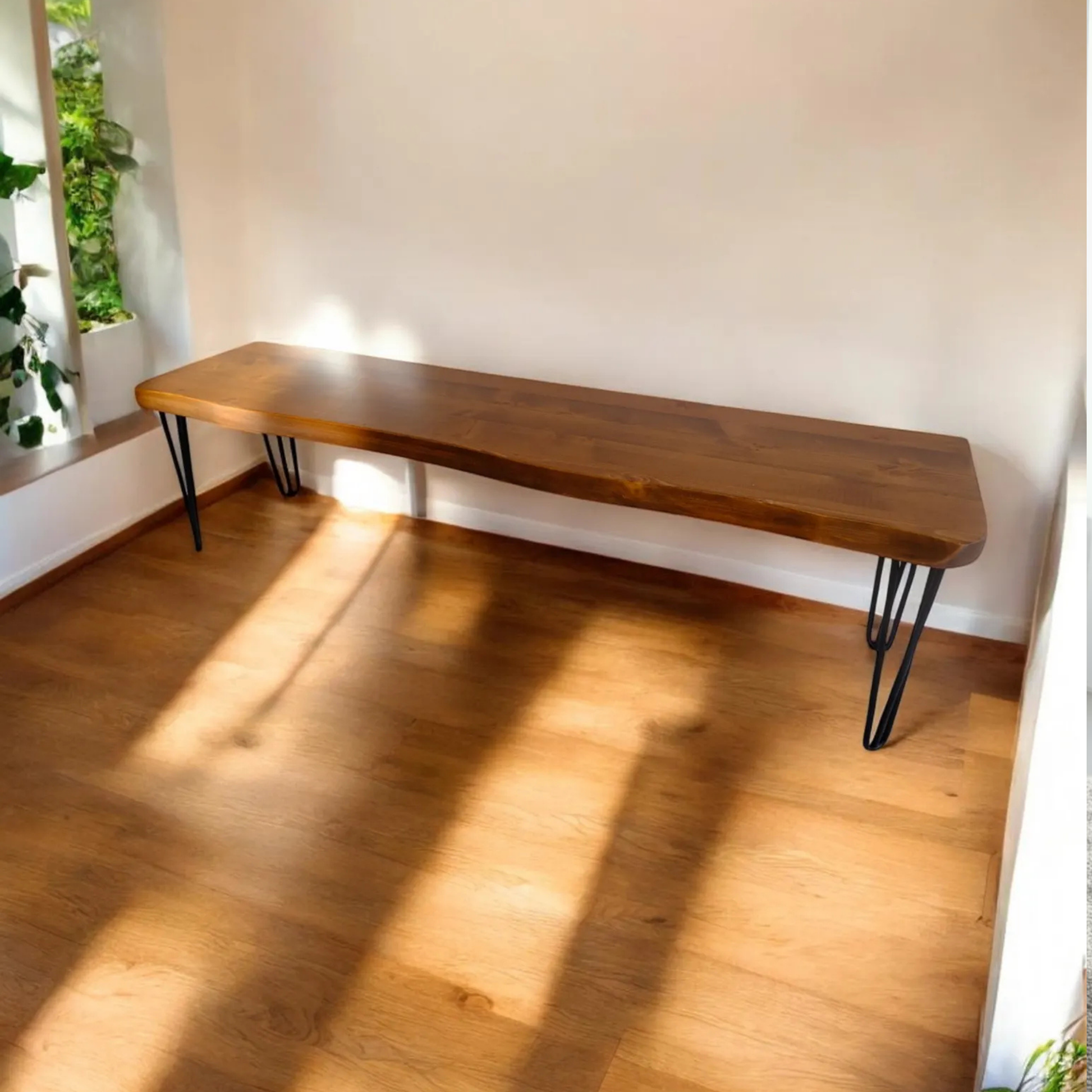 A long live-edge wooden console table with black metal hairpin legs, set against a white wall on a light wood floor with warm sunlight casting shadows.