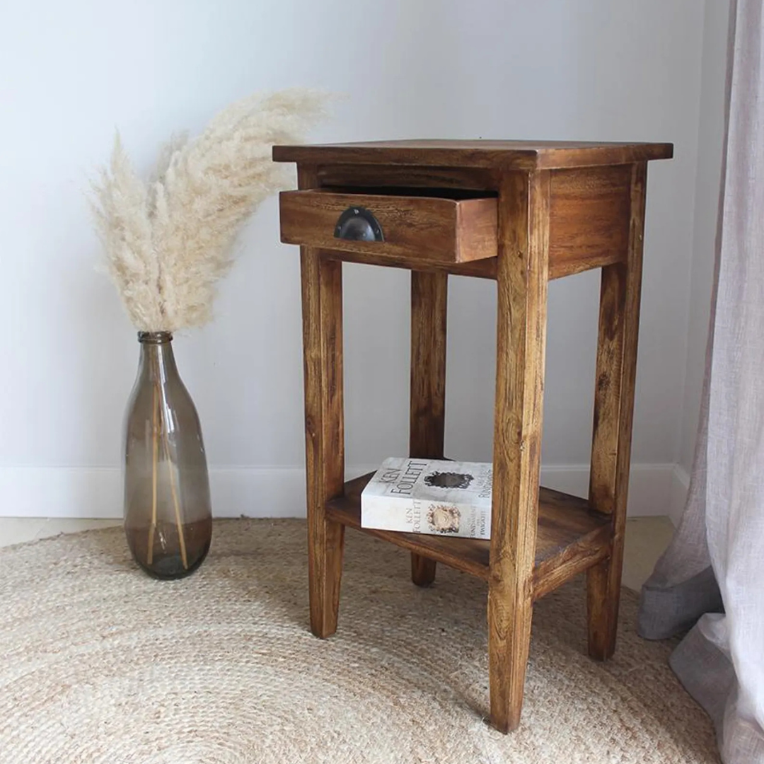 A rustic wooden nightstand with a single drawer and open lower shelf, styled in a room setting next to a glass vase with pampas grass and a book.