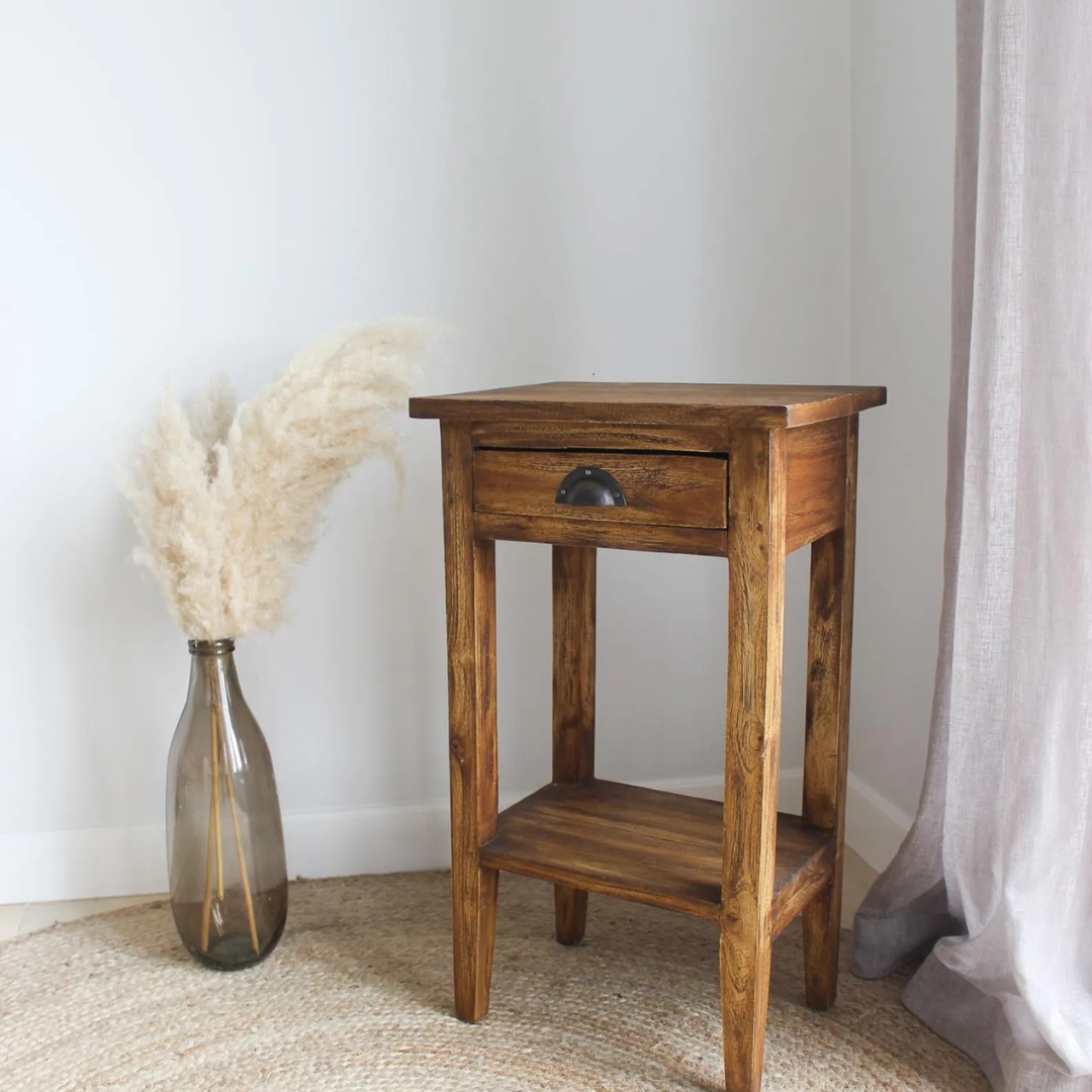 A rustic wooden side table with a single drawer and lower shelf, styled next to a glass vase with pampas grass in a room corner.