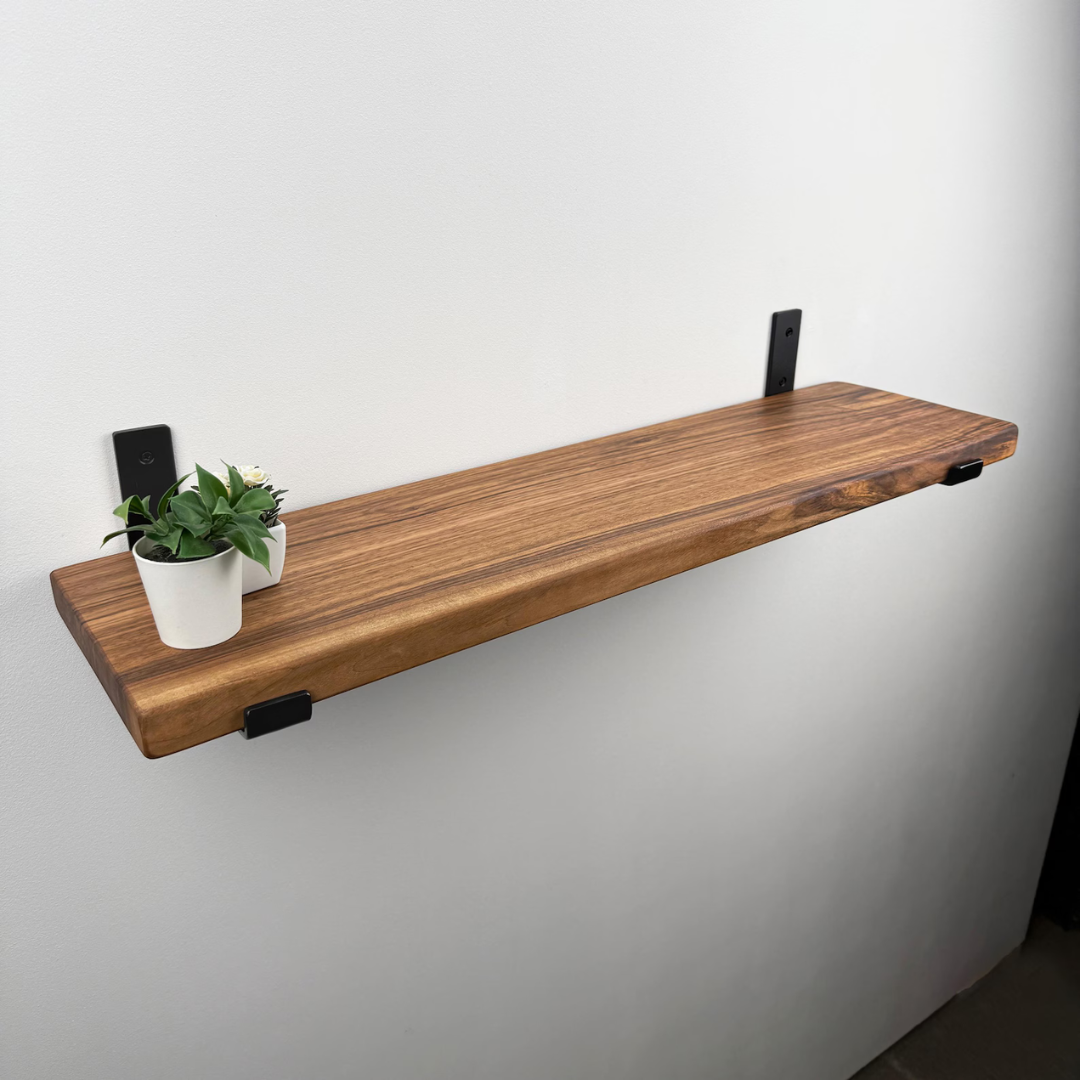 A rustic wooden floating shelf with black metal wall brackets, mounted on a plain white wall and styled with a small potted green plant.