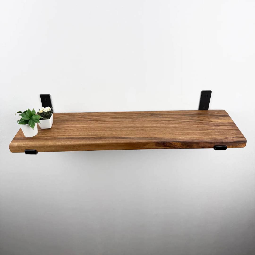 A live-edge wooden floating shelf with black metal brackets, shown mounted on a plain white wall with two small potted plants on the left side.