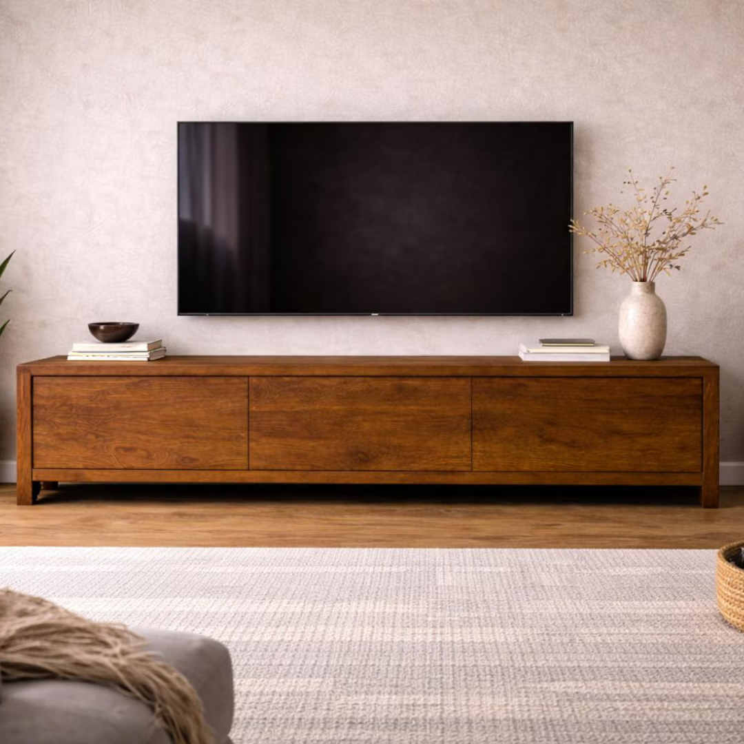 A modern medium-brown wooden media console with three front drawers, styled in a living room setting with a black flat-screen TV mounted on a textured light beige wall above it.