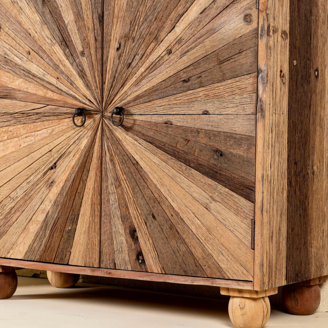 A rustic wooden cabinet with two doors featuring a radial sunburst pattern made from contrasting wood tones, finished with dark metal ring pulls and turned wooden feet.