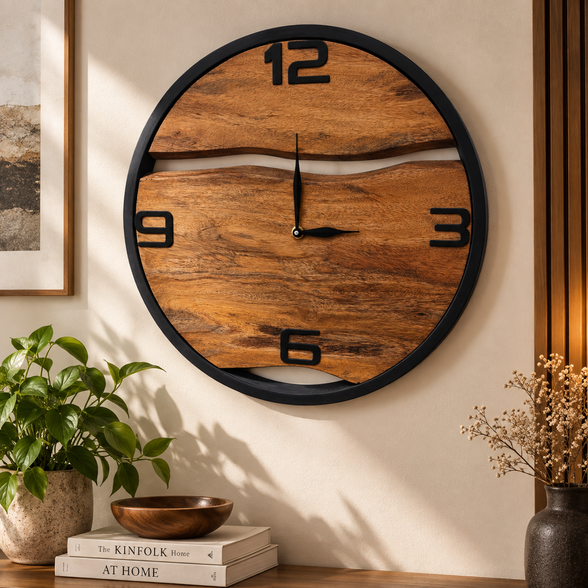 A round wooden wall clock with a live-edge wood grain face and black metal frame, mounted on a beige wall in a styled interior setting with a plant, books, and dried flowers.