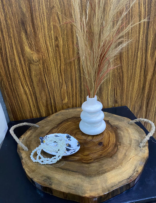 Decorative setup with a white vase, dried grasses, and a wooden board on a dark surface.